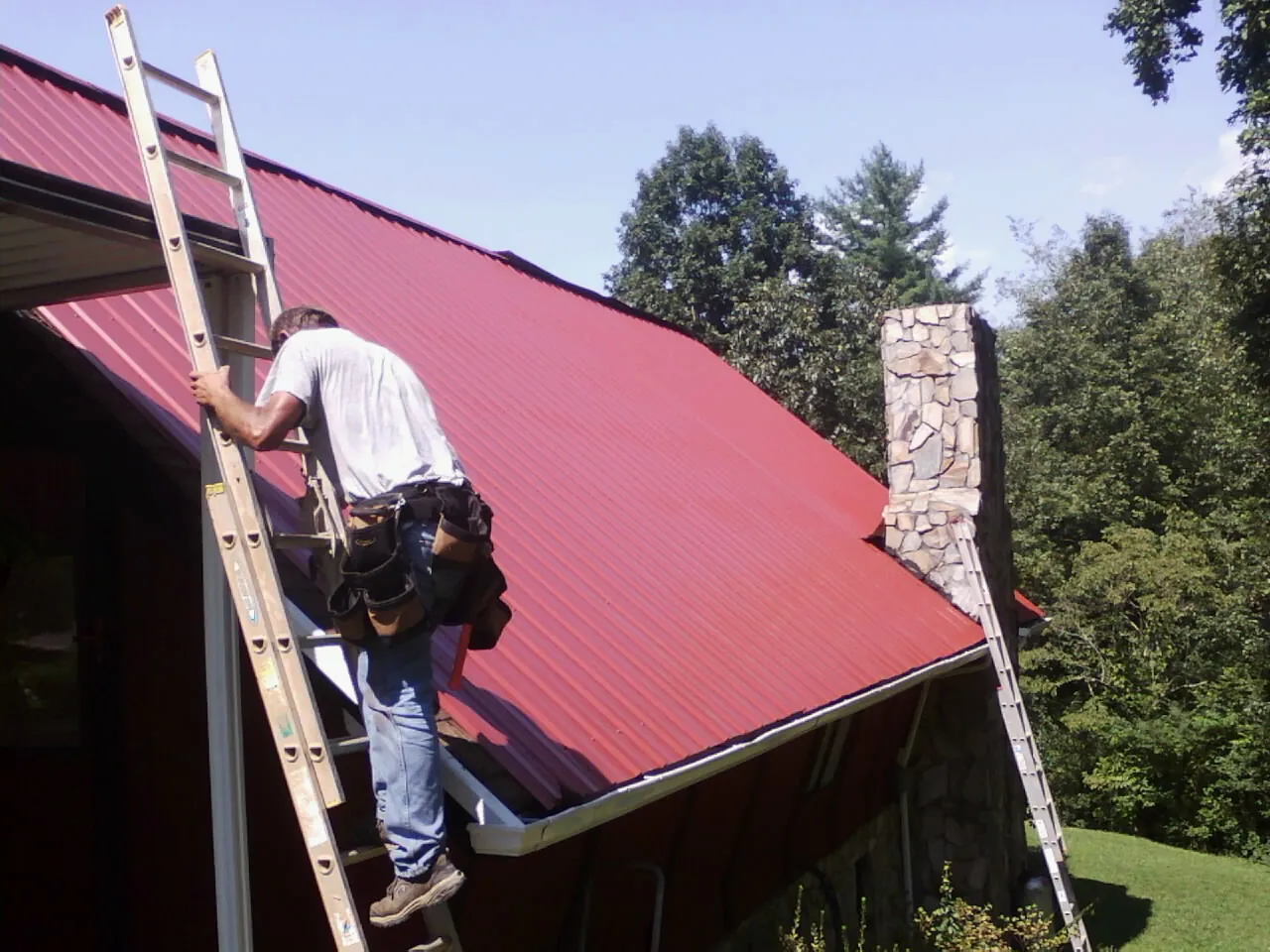 Roofer on a ladder beside a red metal roof and stone chimney — Soffit & Fascia Repair by Roofing Brothers in Coolidge